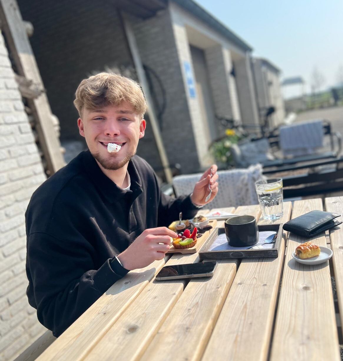 A young man sitting at an outdoor café table, smiling with cream on his face, completely unbothered and enjoying his food.